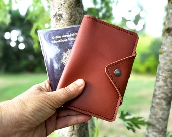 May include: A brown leather passport holder with a flap closure and a metal button. The holder is shown with a French passport inside.