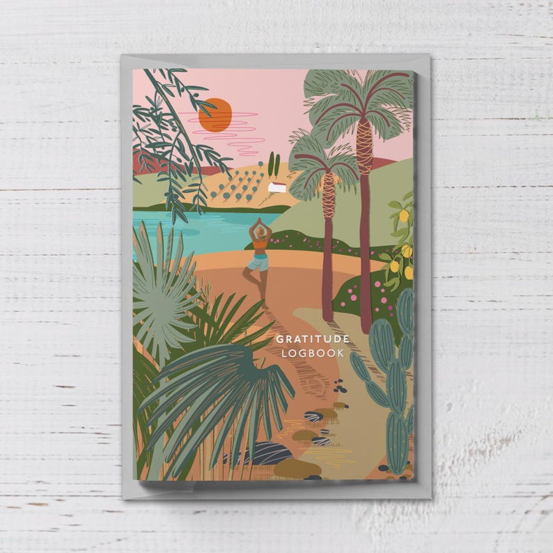 Puede incluir: Un diario con una colorida ilustraci&oacute;n de una escena de playa con palmeras, un lago y una persona haciendo yoga. La portada del diario tiene las palabras "GRATITUDE LOGBOOK" en una fuente sans-serif.