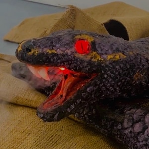 Puede incluir: Primer plano detallado de una cabeza de serpiente con ojos rojos brillantes y boca abierta que revela dientes afilados. La serpiente es de color gris oscuro con detalles amarillos, y la boca brilla en rojo. El fondo incluye un material de yute beige.