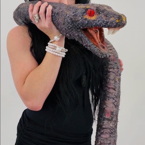 Peut inclure: Un grand serpent gris et noir avec des yeux rouges et une bouche jaune. Le serpent est tenu par une personne portant un haut et un short noirs. La gueule du serpent est ouverte, révélant des dents blanches.