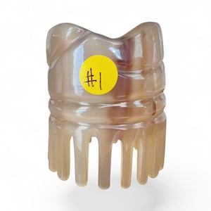 May include: A translucent beige and brown marbled hair claw clip with a smooth, glossy finish. The clip features a rounded top edge and multiple downward-pointing teeth. A circular yellow sticker with the handwritten black text #1 is attached.