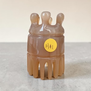 May include: A light brown, carved stone sculpture with a crown-like top and a cylindrical body. The sculpture has a series of vertical supports at the base. A yellow sticker with the text "H#1" is affixed to the body.