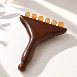 May include: A dark brown wooden massage tool with a T-shaped handle and a rounded knob at the base. The top edge features six small, smooth, translucent amber-colored spherical beads embedded in a row across the wide, flat wooden head.