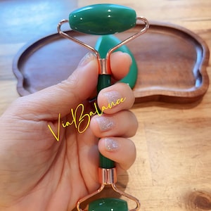 May include: A green jade facial roller with rose gold accents. The roller is held in a hand, showcasing its smooth, polished surface. The design includes two jade stones connected by a rose gold frame and a green handle. A jade gua sha tool is also visible.