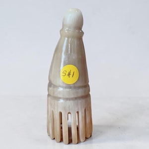 May include: A light beige, carved stone comb with a bottle-shaped handle. The comb has a rounded top and a series of vertical teeth. A yellow sticker with the text "S#1" is affixed to the front.