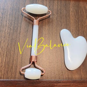May include: A white jade facial roller and a heart-shaped gua sha tool. The roller has a rose gold frame and a white handle. The text "Via Balance" is written in yellow cursive on the wooden surface.
