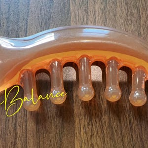 May include: A translucent orange massage tool with a curved design and six rounded protrusions. The text "Via Balance" is written in yellow cursive script. The tool is made of a smooth, glossy material.