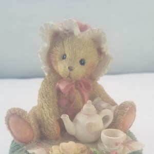 Puede incluir: Un pequeño oso de peluche beige con un gorro blanco con ribete rosa está sentado sobre una base verde de hierba. El oso sostiene una tetera blanca con un diseño de corazón rosa y una cinta rosa. Hay una pequeña taza de té blanca con un diseño de corazón rosa en un platillo junto a la tetera. El oso está sentado sobre una manta de picnic a cuadros blancos y rosas con una galleta amarilla en forma de corazón en un plato.