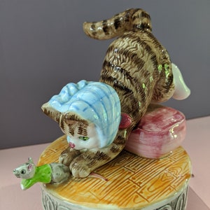 Puede incluir: Una figurita musical de cerámica de un gato atigrado marrón con un gorro azul, abalanzándose sobre un ratón verde y gris. El gato está sentado en un cojín rosa y la base de la figurita está decorada con un suelo de baldosas amarillo y naranja.