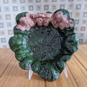 Op de afbeelding: Een groene keramische schaal in de vorm van een blad met een roze bladontwerp rond de rand. De schaal staat op een houten oppervlak.