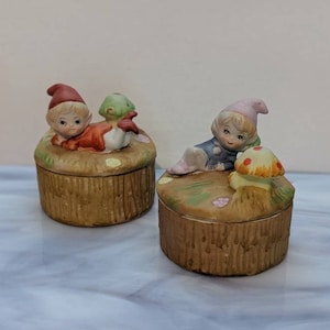 Puede incluir: Dos cajas de cerámica para guardar pequeños objetos con figuritas de duendes. Las cajas son marrones con textura de madera. Un duende lleva un gorro rojo y el otro un gorro morado. Ambos duendes sostienen setas.
