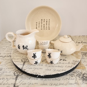 May include: A white ceramic tea set with black calligraphy on the teapot and cups. The set includes a teapot, a pitcher, four cups, and a saucer. The set is on a round wooden tray with a white and black patterned surface.