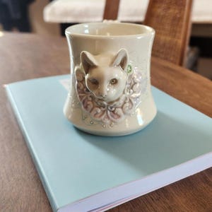 May include: A cream-colored ceramic mug with a sculpted cat face and decorative floral accents. The mug is resting on a light blue notebook with a pink ribbon bookmark. The mug's design features a detailed cat face with green accents.