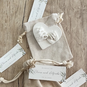 May include: Two small white burlap bags with drawstrings, each containing a white heart-shaped plaster cast of two hands clasped together. Each bag has a white tag attached with the text "Wedding of Olivia and Alfredo" and the date "09/12/2024".