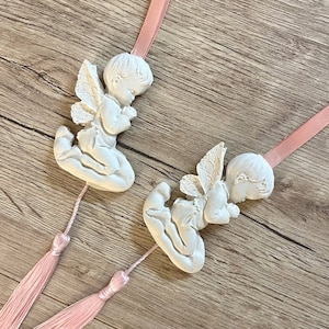 May include: Two white ceramic angel figurines with pink ribbon and tassel accents. The angels are kneeling with their hands clasped in prayer and have wings.