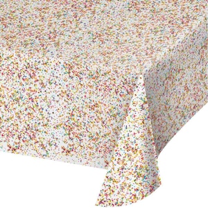 May include: A white rectangular tablecloth covered in a colorful confetti pattern. The confetti is a mix of red, yellow, green, and blue specks. The tablecloth is folded over a table, with the corner of the cloth visible.