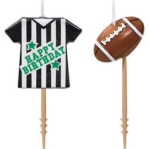 May include: Two birthday cake candles. One is shaped like a black and white striped referee shirt with the text "HAPPY BIRTHDAY" in green. The other is a brown football with white stripes.