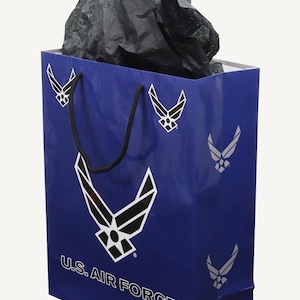 May include: A blue gift bag with the U.S. Air Force logo in white. The bag has black rope handles and is filled with black tissue paper. The words "U.S. AIR FORCE" are printed on the front of the bag.