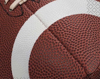 May include: Close-up of a brown football with white stitching and a white stripe. The football has a textured surface and is designed for American football games. The laces are visible at the top of the image.