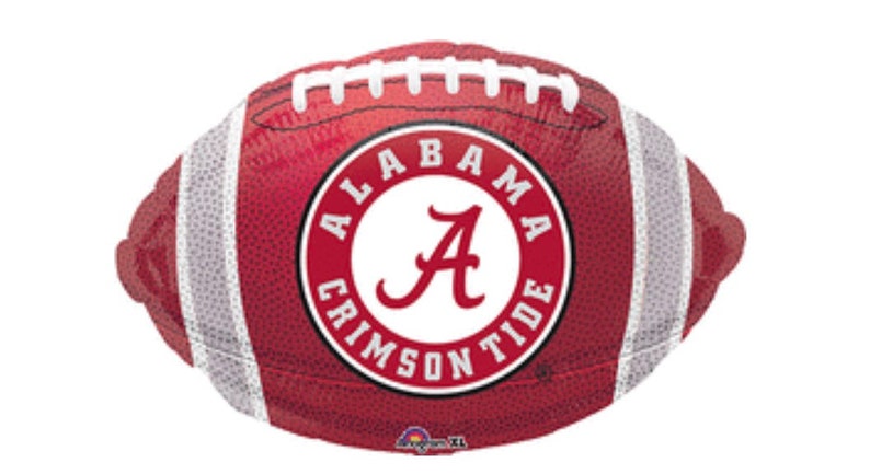 May include: A maroon football-shaped balloon with white stripes and stitching. The center features a circular emblem with the words "ALABAMA CRIMSON TIDE" around a large letter "A".