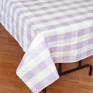 May include: A lavender and white checkered tablecloth draped over a table. The fabric has a classic gingham pattern, with the corner of the cloth folded over, revealing the pattern. The table has black legs and sits on a wooden floor.