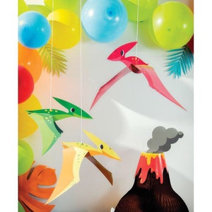 May include: A party scene with colourful balloons and hanging dinosaur decorations. The decorations include pterodactyls in red, green, and yellow, and a volcano. The balloons are in shades of blue, yellow, orange, and green.