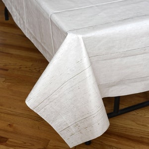May include: A white tablecloth with a wood grain pattern. The tablecloth is draped over a table, with the corner of the cloth folded over. The wood grain pattern is visible on the surface of the tablecloth.