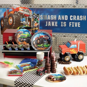 May include: A festive monster truck-themed birthday party setup. Includes plates, napkins, cups, and a decorative red truck. The backdrop features a monster truck graphic and the text "SMASH AND CRASH JAKE IS FIVE".