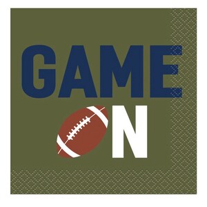 May include: A square paper napkin with a football theme. The words "GAME ON" are printed in navy blue and white, with a brown and white football graphic in the center. The background is olive green.
