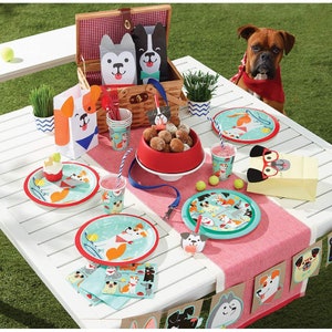 May include: A festive picnic scene with dog-themed party supplies. Includes plates, cups, napkins, and treat bags featuring playful dog illustrations. A picnic basket, dog-shaped decorations, and a red dog bowl add to the theme.