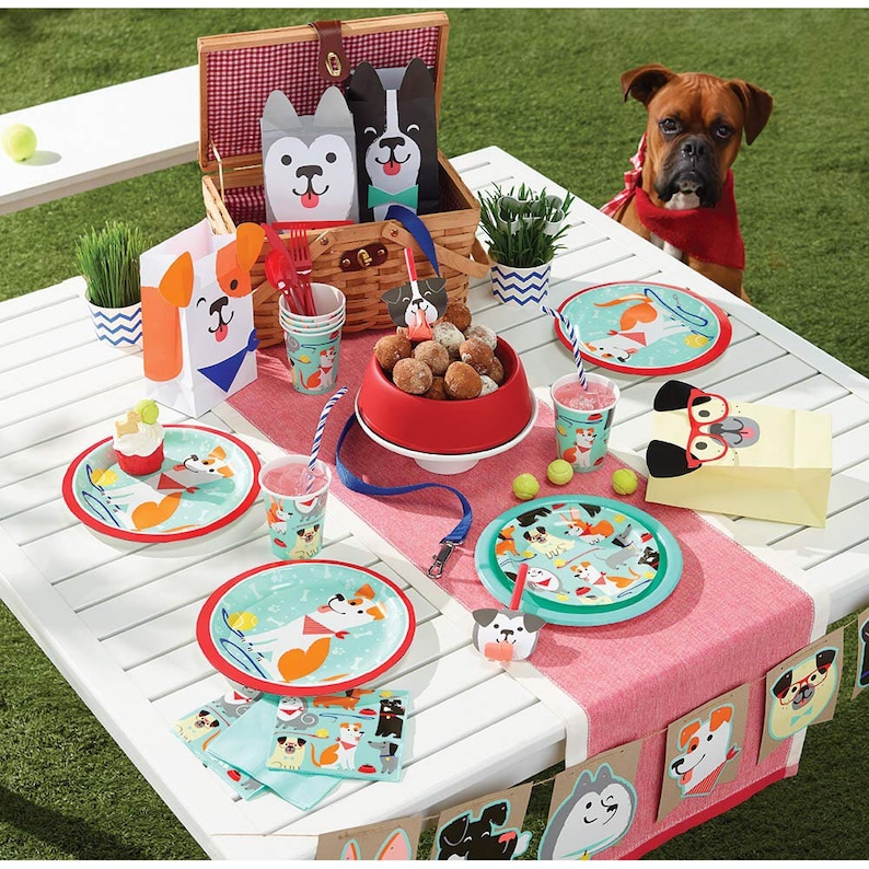 May include: A festive picnic scene with dog-themed party supplies. Includes plates, cups, napkins, and treat bags featuring playful dog illustrations. A picnic basket, dog-shaped decorations, and a red dog bowl add to the theme.