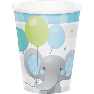 May include: A paper cup with a baby elephant design. The cup features a gray elephant holding green and blue balloons. The background has blue polka dots and stripes. The rim of the cup is white.