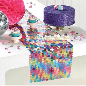 May include: A festive party table setting with a colorful sequin table runner. A purple glitter cake sits on a silver stand, surrounded by cupcakes and decorative accents. A pink honeycomb ball and disco ball add to the celebratory atmosphere.