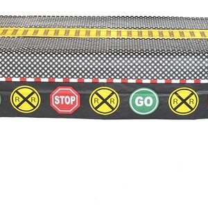 May include: A rectangular black and white checkered tablecloth with a train track design. The border features railroad crossing signs, stop signs, and go signs. A yellow stripe runs down the center of the tablecloth.
