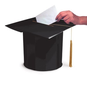 May include: A black graduation cap-shaped ballot box with a gold tassel. A hand is inserting a white paper ballot into the slot on top. The ballot box is on a white background.