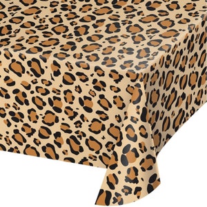 May include: A tan tablecloth with a leopard print pattern. The pattern features brown and black spots of varying sizes and shapes, creating a wild animal print design. The tablecloth is folded over a table corner.