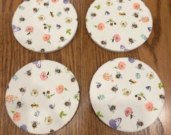 May include: Four round white coasters with a floral and bee pattern. The coasters feature small pink, purple, and yellow flowers, green leaves, and small brown bees. There is also a larger purple butterfly on each coaster.