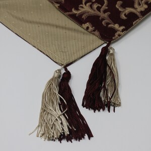 May include: Two tassels, one burgundy and one cream, hang from a burgundy and cream patterned table runner.