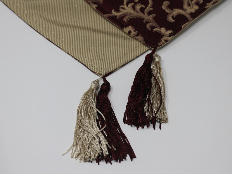May include: Two tassels, one burgundy and one cream, hang from a burgundy and cream patterned table runner.