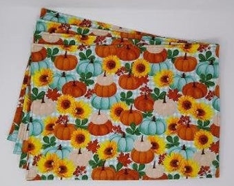 May include: Set of four rectangular placemats with a repeating pattern of pumpkins, sunflowers, and fall foliage in orange, teal, yellow, and cream colors. The placemats are stacked on a white surface.