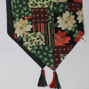 May include: A black and green table runner with a patchwork design featuring red and white poinsettia flowers. The runner has two tassels at the ends.