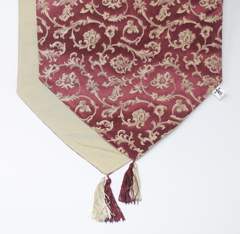 May include: A burgundy table runner with a gold floral pattern and tassels. The runner has a beige border and a small tag with the text "La Table" on it.