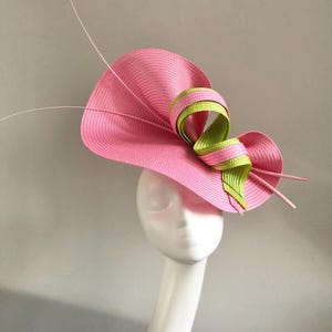 Op de afbeelding: Een roze zonnehoed met een brede rand en een decoratieve strik. De strik is gemaakt van groen en roze lint. De hoed wordt getoond op een witte mannequinhoofd. Dunne roze en witte accenten steken uit de hoed.