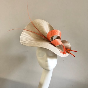 May include: A beige straw hat with a wide brim, adorned with a coral-colored bow and slender orange accents. The hat is displayed on a white mannequin head, showcasing its elegant design and craftsmanship. This hat is perfect for special occasions.