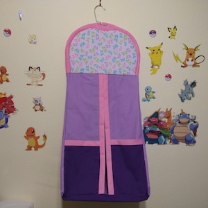 May include: A purple and pink nursery organizer hanging on a wall. The organizer has a pocket design with a pink trim and a top section with a pink border and a butterfly pattern. The wall is decorated with various Pokemon character stickers.