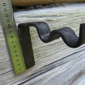 Op de afbeelding: Een zwarte metalen gordijnroedebeugel met een golvend ontwerp. De beugel is met twee schroeven aan een houten oppervlak bevestigd. Een liniaal is zichtbaar op de afbeelding, wat aangeeft dat de beugel ongeveer 10 cm lang is.