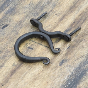 May include: A hand-forged, black iron bracket with two wood screws. The bracket has a curved, circular design with decorative scrollwork at the ends. The metal has a textured, rustic appearance, suggesting it is handmade.