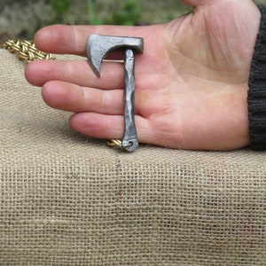 May include: A small, silver-colored axe keychain with a textured handle. The axe is attached to a gold chain.