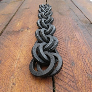 May include: A dark grey, hand-forged metal decorative chain made of interlocking rings. The chain is displayed on a rustic, weathered wooden surface with visible grain and knots. The metal has a textured, industrial appearance.