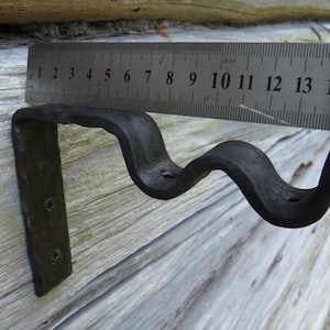 Op de afbeelding: Een zwarte metalen beugel met een golvend ontwerp, gemeten met een liniaal die in centimeters is gemarkeerd. De beugel is bevestigd aan een houten oppervlak.
